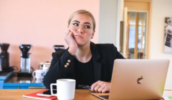 A woman looks up in thought, unamused, at a desk with a mug and laptop, in a bright pink room